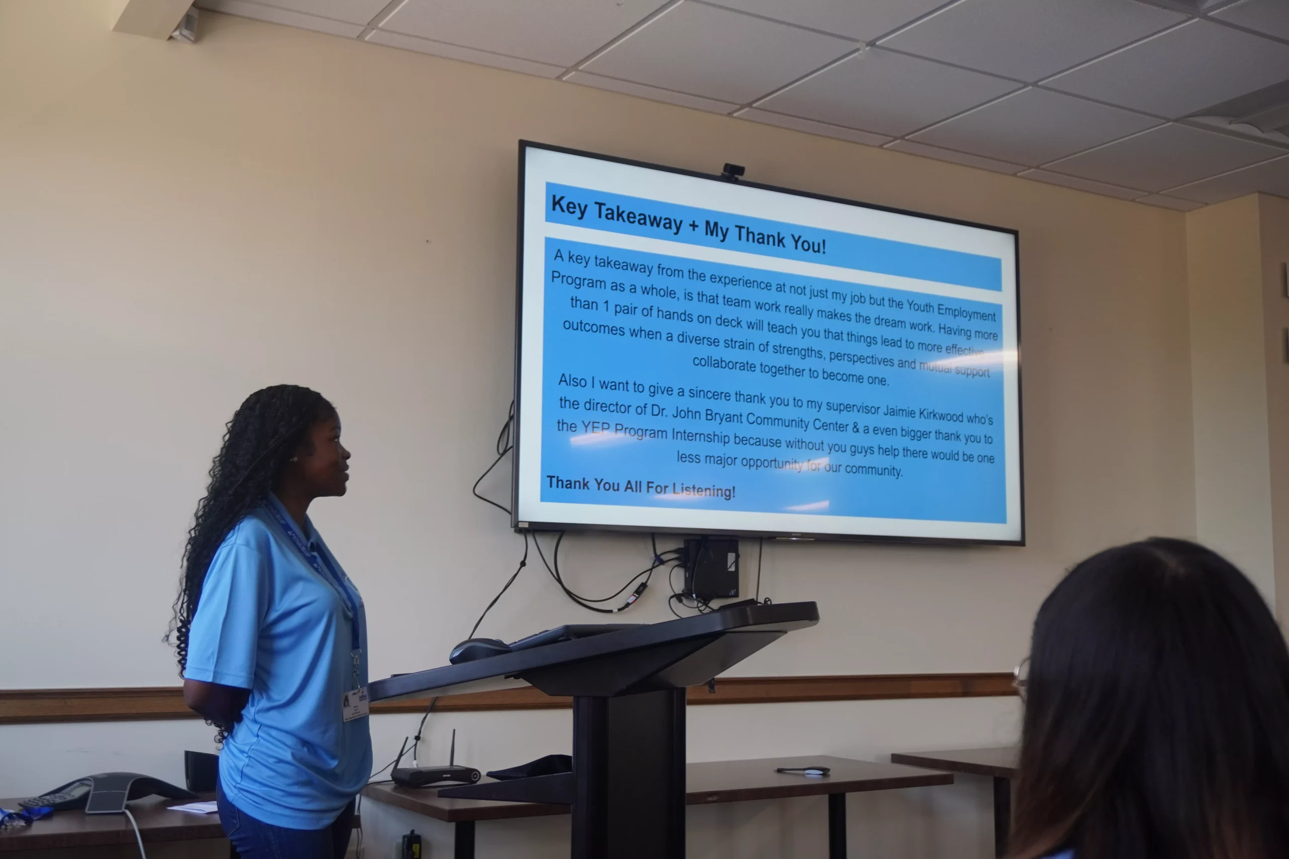 Presenter stands near a screen displaying "Key Takeaways + My Thank You!" with text about program support.