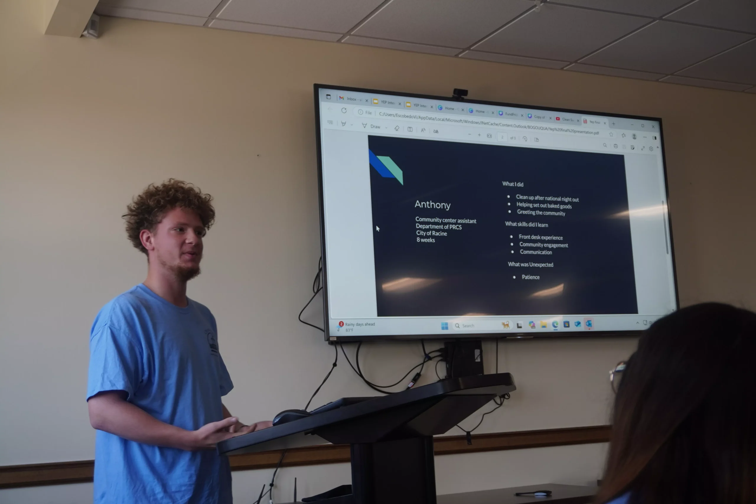 Man presenting information on a large screen during a presentation. The screen displays text and a logo.