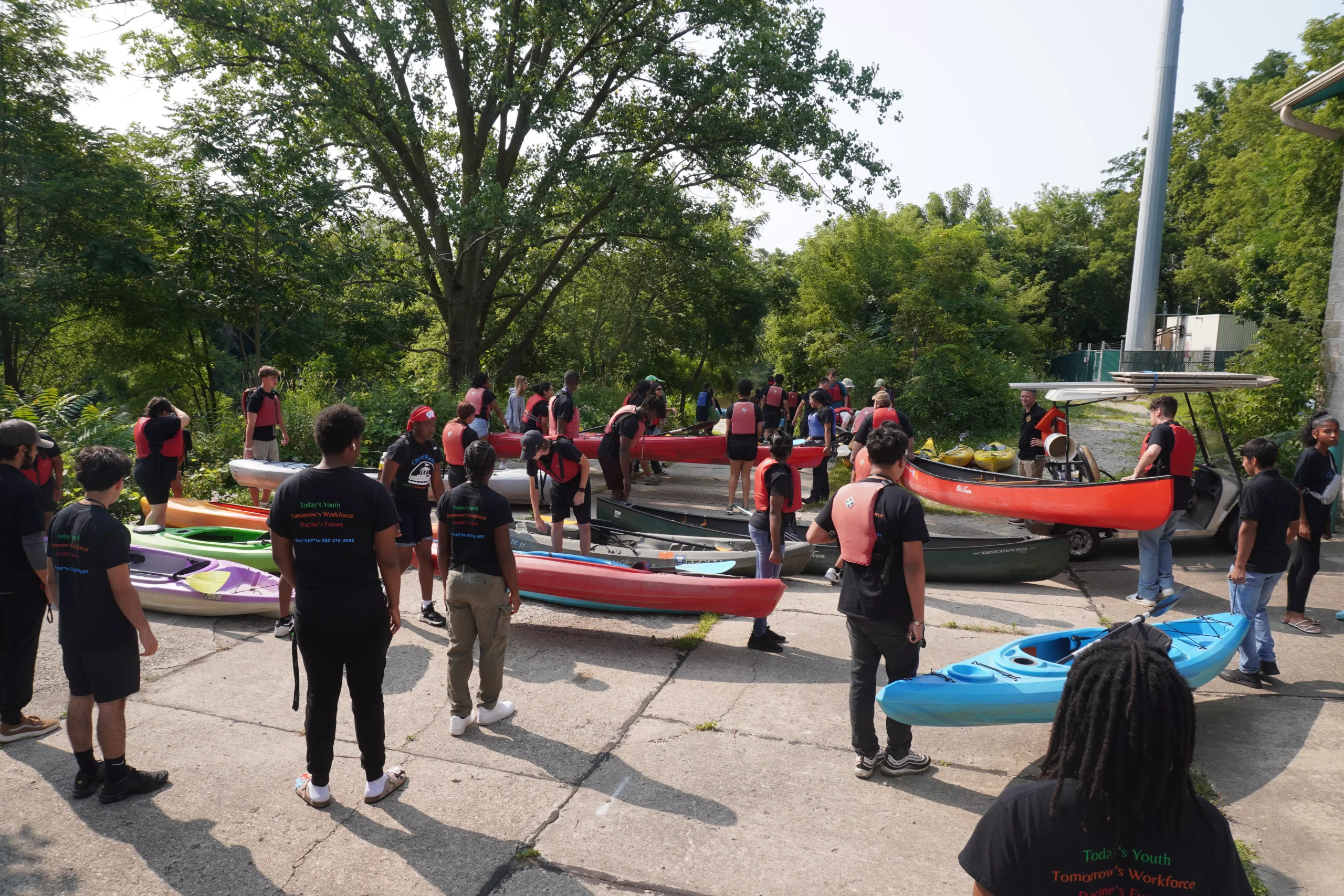 Group of young people wearing life vests stand near kayaks and canoes outdoors, preparing for water activity.