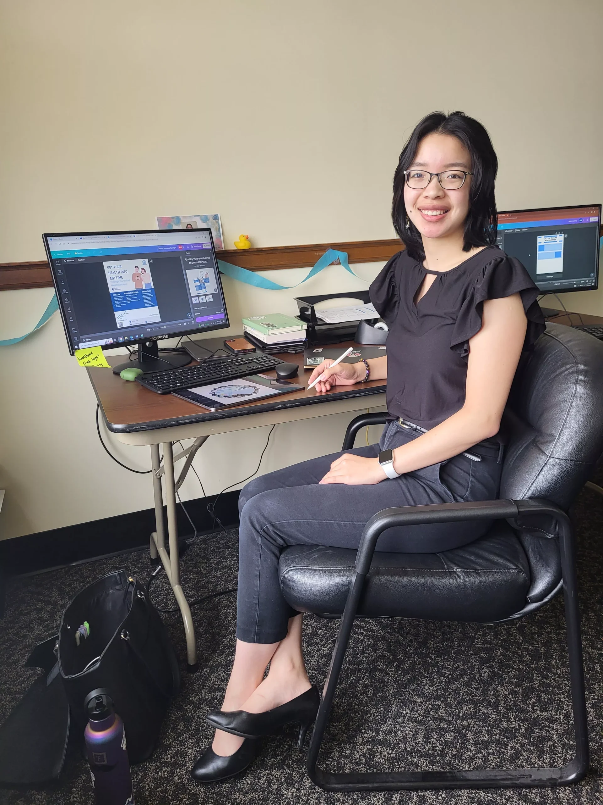 Young woman with glasses working at a computer with a graphic design program open, smiling at the camera.