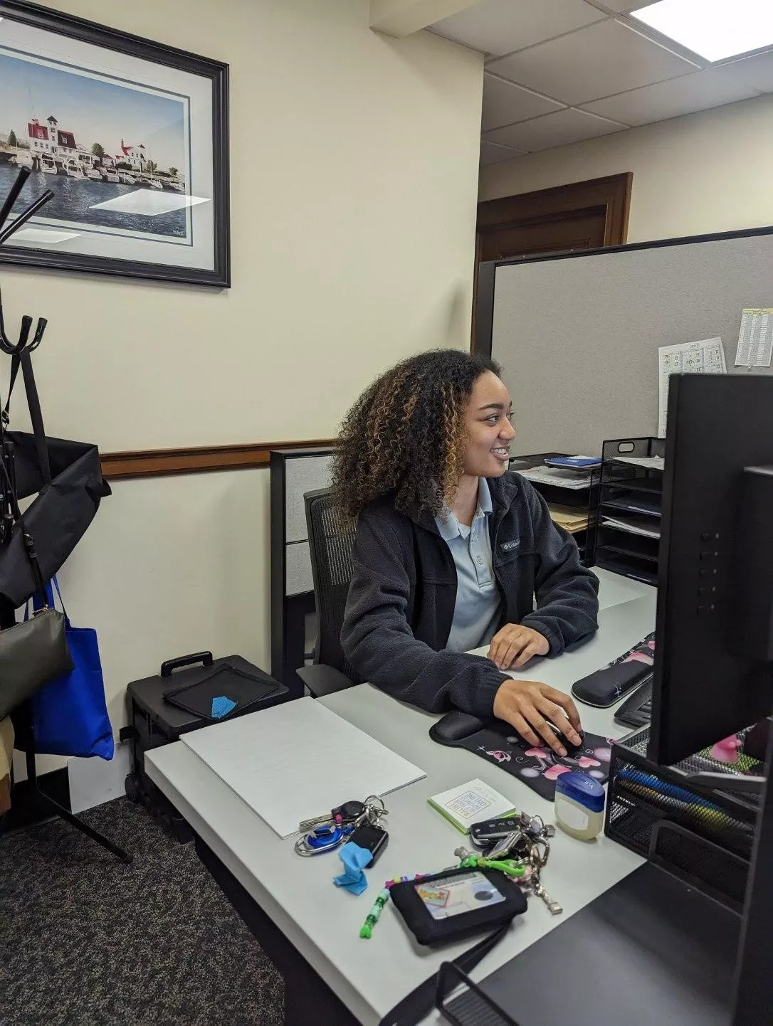 Woman with curly hair works at her desk using a computer in an office setting. Artwork hangs on the wall.