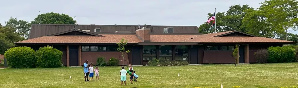Children play on the lawn in front of a one-story building with a brown roof and brick chimney.